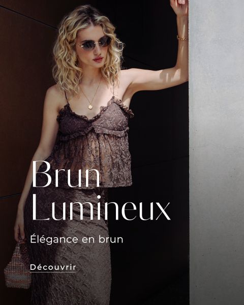 Une femme vêtue d'une robe en dentelle marron et portant des lunettes de soleil se tient au soleil devant un mur. Le texte dit : "Brun Lumineux, Élégance en brun, Découvrir."