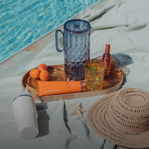 Un plateau au bord de la piscine avec de la verrerie, des abricots, de la crème solaire, un vaporisateur, un chapeau de soleil, une tasse blanche et une étoile de mer sur une serviette vert clair.
