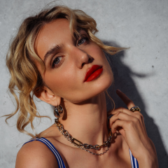 Une femme aux cheveux blonds ondulés, au rouge à lèvres rouge vif et aux bijoux en or pose devant un mur de béton, touchant sa nuque et regardant l'appareil photo avec assurance.