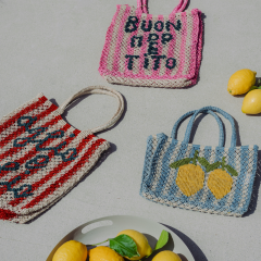 Trois sacs tissés colorés, deux avec du texte italien et un avec des citrons, sont posés sur une surface claire à côté d'une assiette de citrons et de basilic.