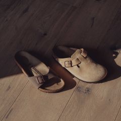 Deux sandales Birkenstock dépareillées - l'une à bout ouvert et l'autre à bout fermé - avec des boucles sur un plancher en bois ensoleillé.