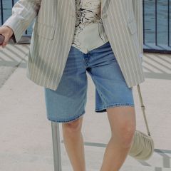 Une personne, portant un blazer à rayures claires, une blouse imprimée et un short en jean, se tient à l'extérieur près d'une balustrade et tient un petit sac à main beige.
