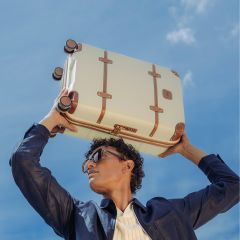 Une personne avec des lunettes de soleil et une veste sombre tient une valise couleur crème avec des sangles marron au-dessus de sa tête devant un ciel bleu.