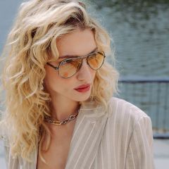 Une femme aux cheveux blonds bouclés, portant des lunettes de soleil aviateur teintées, un blazer beige rayé et un collier chaîne épais, se tient au bord d'un plan d'eau.