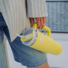 Personne en short en jean et veste rayée, tenant un grand gobelet Stanley jaune avec paille et anse.