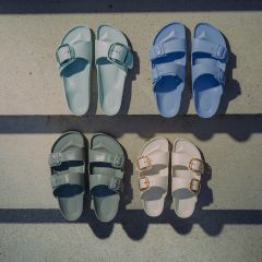 Quatre paires de sandales en bleu pastel, vert pastel, vert foncé et blanc cassé, disposées sur une surface en béton et traversées par des ombres.