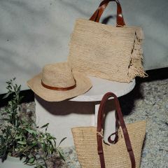 Chapeau de paille tressé et deux sacs de paille avec des poignées marron sur une table ronde blanche à côté d'une plante en pot verte, en extérieur sous le soleil.
