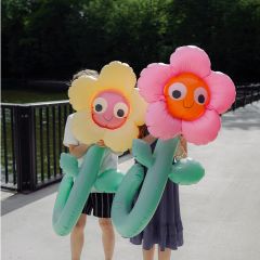 Deux personnes tiennent de grandes fleurs gonflables jaunes et roses devant leur visage, tandis qu'elles se tiennent sur un chemin ensoleillé près d'une balustrade et de plantes vertes.