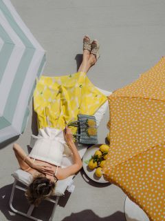 Une femme en jupe jaune se détend sur une chaise longue sous des parasols par une journée ensoleillée, avec des citrons et un sac au motif de citrons à ses côtés.