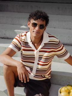Un homme aux cheveux bouclés et aux lunettes de soleil est assis sur un escalier extérieur et porte une chemise rayée à manches courtes ; à côté de lui se trouve un bol de citrons.
