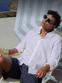 Un homme avec des lunettes de soleil et une chemise blanche se détend sur une chaise longue, une boisson au citron à la main, sous un parasol rayé par une journée ensoleillée.