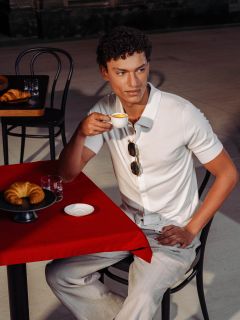 Un homme en chemise blanche est assis à une table rouge, buvant un expresso, avec un croissant et un verre d'eau devant lui dans un café en plein air.