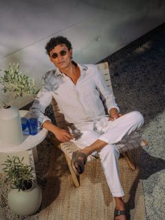 Un homme avec des lunettes de soleil et des vêtements blancs se détend sur une chaise à l'extérieur, à côté d'une petite table avec des verres bleus et un vase, sur un tapis tissé.