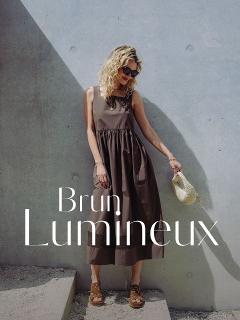 Une femme vêtue d'une robe marron et portant des lunettes de soleil se tient au soleil devant un mur en béton, tenant un chapeau de paille à la main. Le texte indique : « Brun Lumineux ».
