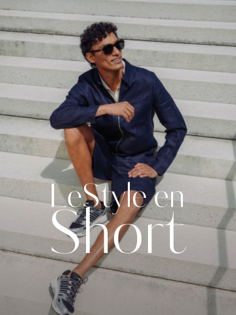 Un homme avec des lunettes de soleil et un costume-short bleu marine est assis en souriant sur un escalier extérieur. Sur l'image, il est écrit : « LeStyle en Short. ».