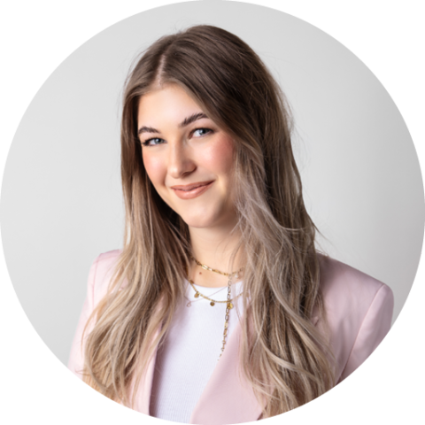 Souriante jeune femme aux longs cheveux châtains clairs, portant un blazer rose et un haut blanc, et se tenant devant un fond gris clair.