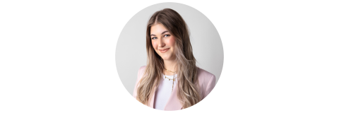 Souriez femme aux longs cheveux châtains clairs, vêtue d'un blazer rose et de colliers superposés, photographiée sur un fond gris clair.