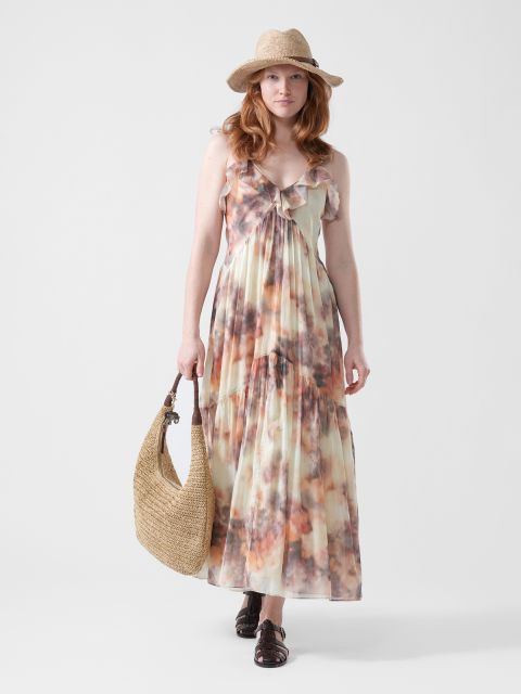 Une femme, vêtue d'une maxi-robe fluide et colorée, d'un chapeau de paille et de sandales, tenant un grand sac tissé, se tient devant un fond blanc uni.