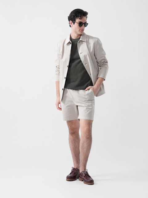 Un homme portant des lunettes de soleil, une veste beige, une chemise vert olive, un short beige et des chaussures marron se tient devant un arrière-plan blanc uni.