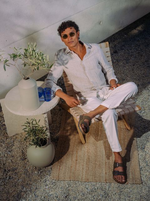 Un homme avec des lunettes de soleil et des vêtements légers se détend sur une chaise longue à côté d'une petite table avec des plantes et des verres, à l'extérieur sur un sol en galets et un tapis tissé.
