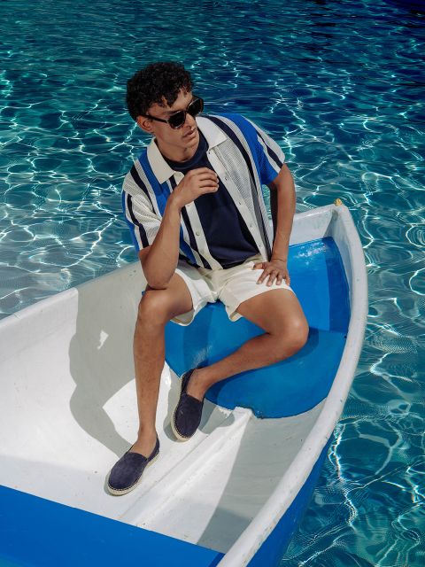 Un homme avec des lunettes de soleil, une chemise rayée et un short est assis détendu dans un petit bateau blanc et bleu sur l'eau bleue claire et scintillante.