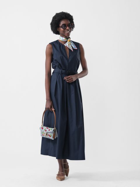 Femme dans une robe bleu marine sans manches, un foulard coloré, des lunettes de soleil, un sac à main à motifs et des chaussures marron, devant un fond blanc.