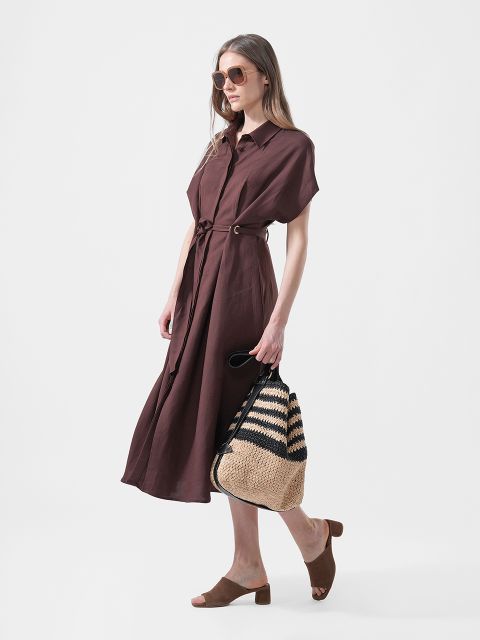 Une femme porte une robe midi marron avec une ceinture, des sandales coulissantes marron, des lunettes de soleil et un sac cabas en paille tressée avec des rayures noires.