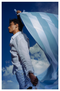 Personne en chemise blanche, tenant un tissu rayé bleu et blanc, devant un ciel bleu avec des nuages.