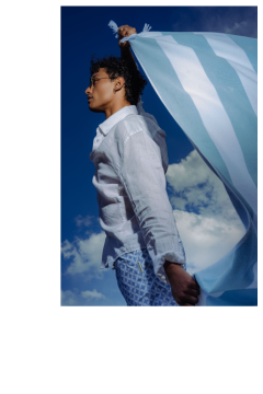 Une personne en chemise blanche et short à motifs tient un tissu rayé bleu et blanc devant un ciel bleu avec des nuages.