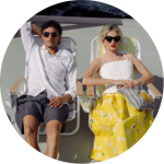 Ein Mann und eine Frau sitzen auf Liegestühlen in der Sonne. Der Mann trägt ein weißes Hemd und kurze Hosen, die Frau eine Sonnenbrille und einen gelb gemusterten Rock.