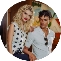 Une femme aux cheveux blonds et bouclés et un homme aux cheveux courts et bouclés posent ensemble dans une maison ; elle sourit, tandis qu'il regarde l'appareil photo avec des lunettes de soleil sur sa chemise.