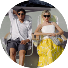 Un homme et une femme sont assis sur des chaises longues au soleil. L'homme porte une chemise blanche et un short, la femme des lunettes de soleil et une jupe à motifs jaunes.