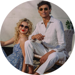 Deux personnes avec des lunettes de soleil se détendent en plein air ; l'une est assise sur une chaise, l'autre est assise à côté, toutes deux sourient et portent des vêtements légers et estivaux.