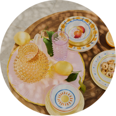 Assiettes colorées à motifs de fruits, verres roses et violets et fruits jaunes sur une table ronde en bois avec un set de table rose.