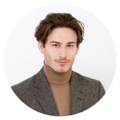 Jeune homme aux cheveux courts et bruns, portant un blazer gris et un pull à col roulé marron clair, et posant devant un fond clair et uni.