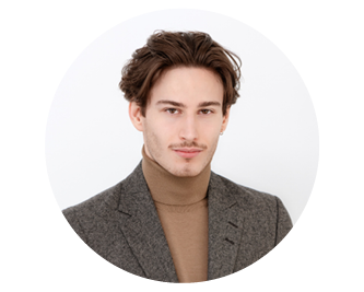 Jeune homme aux cheveux courts et bruns, portant un blazer gris et un pull à col roulé marron clair, et posant devant un fond clair et uni.