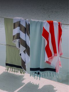 Quatre serviettes de différentes couleurs et motifs sont suspendues à une clôture en corde au bord de l'eau, projetant des ombres sur le sol en dessous.