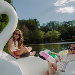 Deux enfants avec des lunettes de soleil se détendent sur un pédalo en forme de cygne sur un lac, entouré d'arbres sous un ciel ensoleillé.