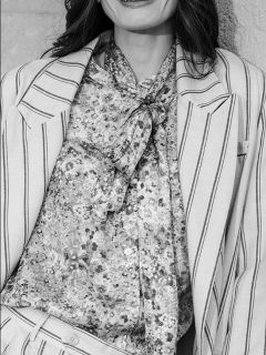 Une femme dans un tailleur rayé avec un chemisier à motifs et un nœud papillon, souriante ; la photo est en noir et blanc et la montre des épaules à la taille.