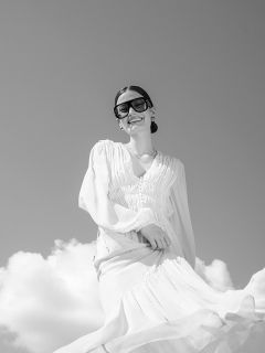 Une femme vêtue d'une robe blanche fluide et de grandes lunettes de soleil sourit sous un ciel nuageux ; la photo est en noir et blanc.