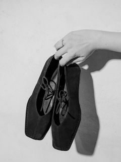 Une main avec une bague au doigt tient une paire de chaussures à lacets noires à bout carré devant un fond clair et uni.