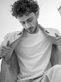 Photo en noir et blanc d'un homme aux cheveux bouclés qui regarde vers le bas ; il porte une veste légère par-dessus un simple t-shirt blanc et tient la veste ouverte.