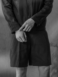 Photo en noir et blanc d'une personne en chemise et short, les mains croisées devant la taille, debout devant un mur texturé.