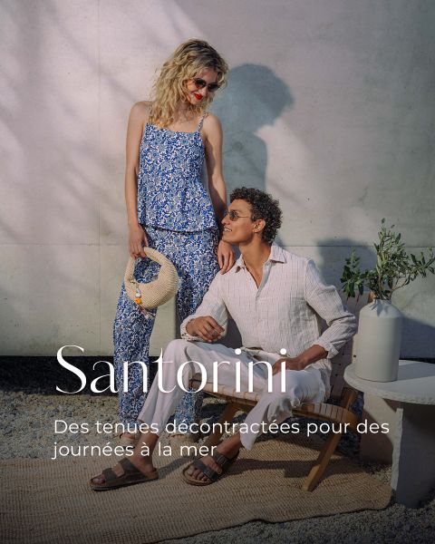 Une femme dans une tenue bleue à fleurs et un homme en blanc sont assis devant un mur ensoleillé et font la promotion de vêtements décontractés. Texte : "Santorini. Des tenues décontractées pour la mer.