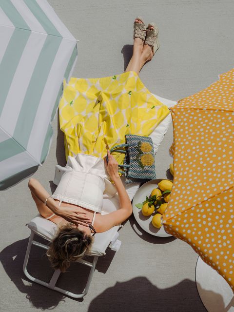 Une femme en jupe jaune est assise sur une chaise sous des parasols, avec des citrons dans un bol et un sac à proximité sur une terrasse ensoleillée.