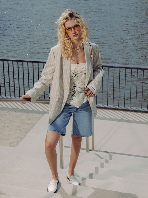 Une femme qui porte des lunettes de soleil, un blazer rayé, un haut fleuri, un short en jean et des chaussures blanches, se tient sur une promenade au bord de l'eau avec une balustrade derrière elle.