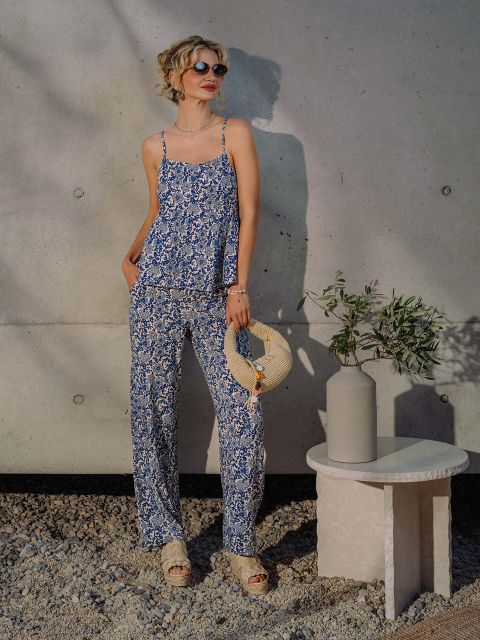 Une femme en haut fleuri bleu et pantalon assorti, portant un chapeau de paille, se tient à une table avec un vase et une branche d'olivier, devant un mur en béton.