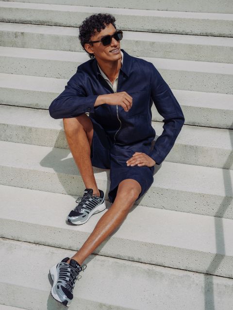Un homme en short et veste bleu marine, lunettes de soleil et baskets, est assis sur un escalier en béton extérieur, souriant et se relaxant sous un soleil éclatant.