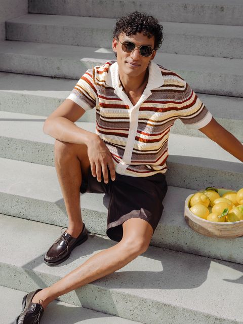 Un homme avec des lunettes de soleil et une chemise rayée est assis sur un escalier extérieur à côté d'un panier de citrons et sourit décontracté au soleil.