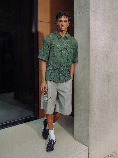 Un homme, portant une chemise verte à manches courtes, un short gris, des chaussettes blanches et des chaussures basses noires, est appuyé contre un mur et tient des lunettes de soleil dans une main.